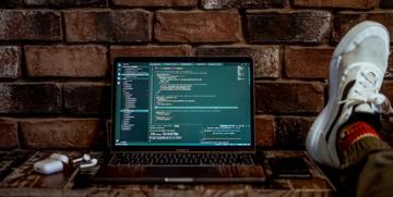 MacBook Pro open on a shelf against a brick wall. The active application is Visual Studio Code with open javascript files.
